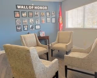 A small seating area with four beige upholstered armchairs arranged around a dark wooden side table. The room has light-colored walls and a large window with blinds letting in natural light. On one wall, there is a display titled 'WALL OF HONOR' featuring framed photos and memorabilia.