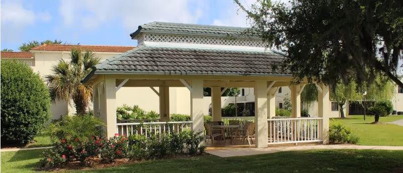 Outdoor gazebo structure with a tiled roof and white railings, surrounded by green grass, bushes, and trees, with a few chairs and a table inside the gazebo. In the background, there is a light-colored building with a red-tiled roof and palm trees nearby.