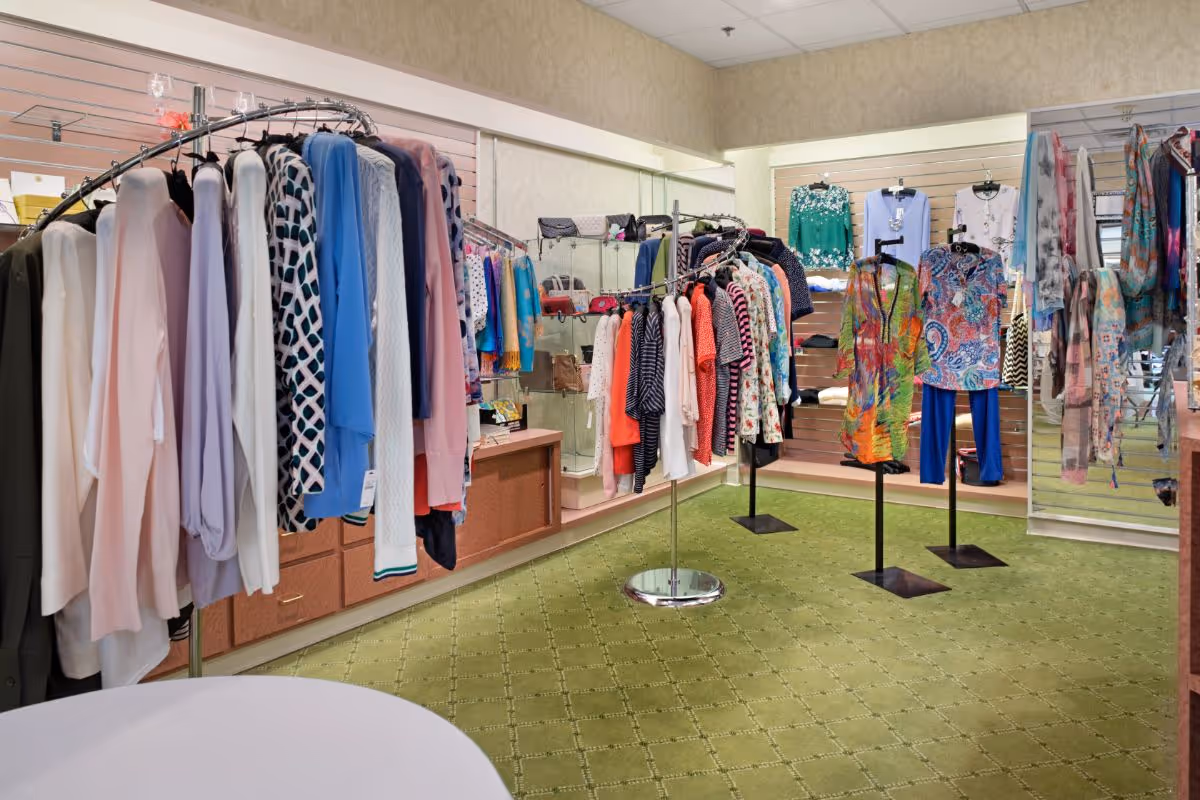Interior view of a clothing boutique area within a facility, featuring racks and mannequins displaying various colorful women's clothing items such as sweaters, blouses, and scarves. The floor is covered with green carpet and the walls have light-colored wallpaper with shelves and display cases holding handbags and accessories.