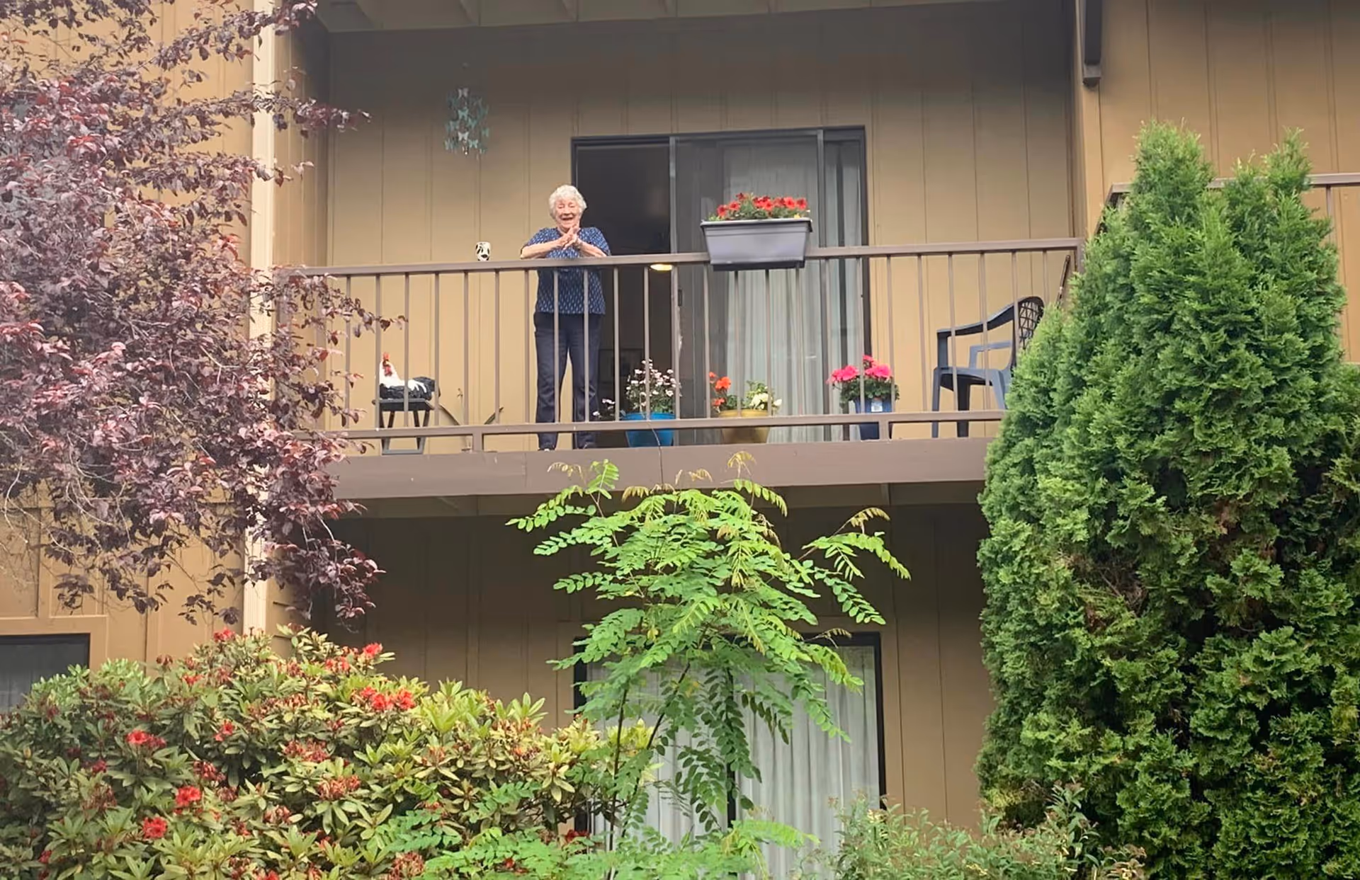An elderly woman stands on a second-floor balcony of a brown apartment building with potted flowers and surrounding trees and shrubs.