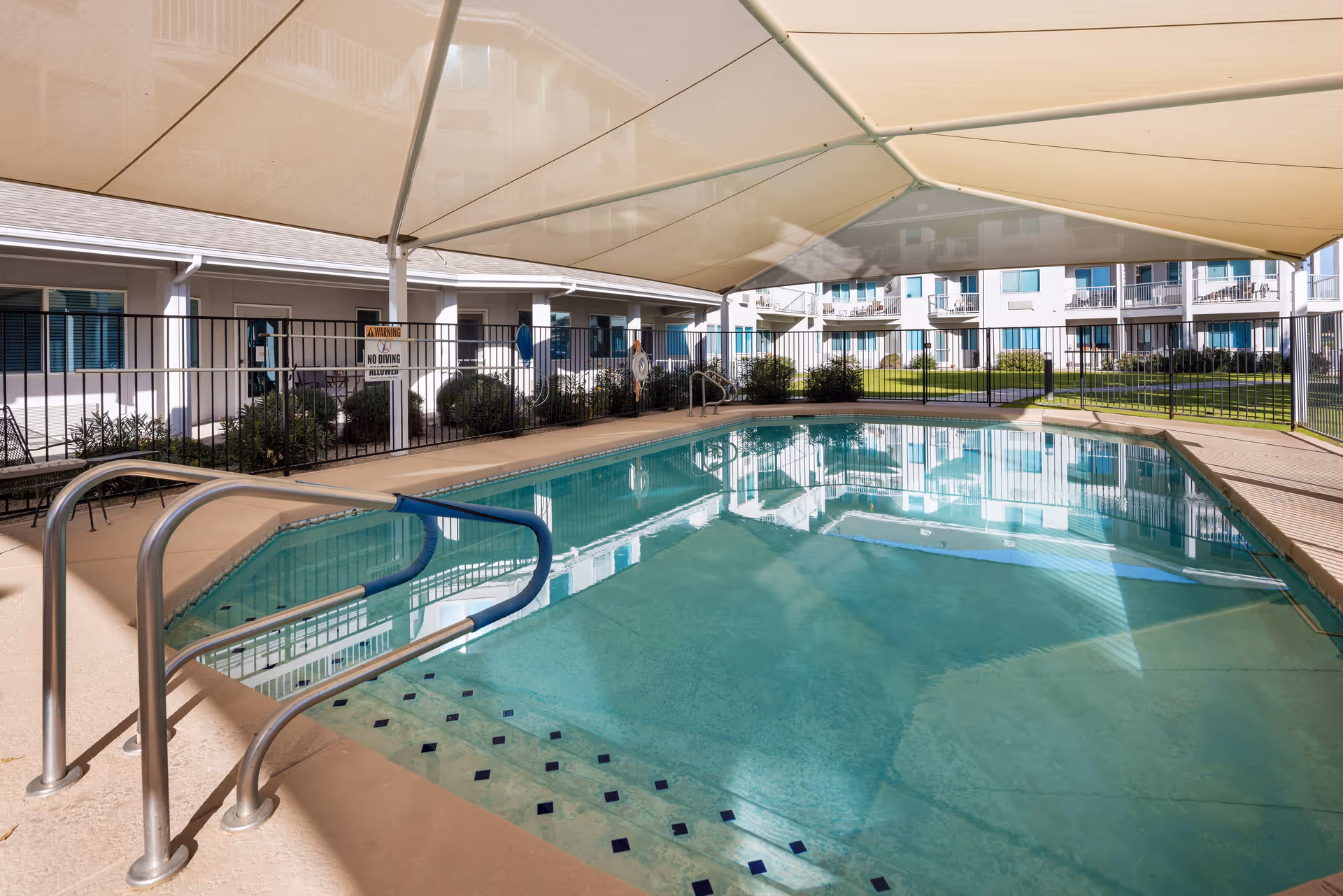 Outdoor swimming pool with a shaded canopy overhead, surrounded by a safety fence. In the background, there are multi-story residential buildings with balconies and windows, and some greenery around the pool area.
