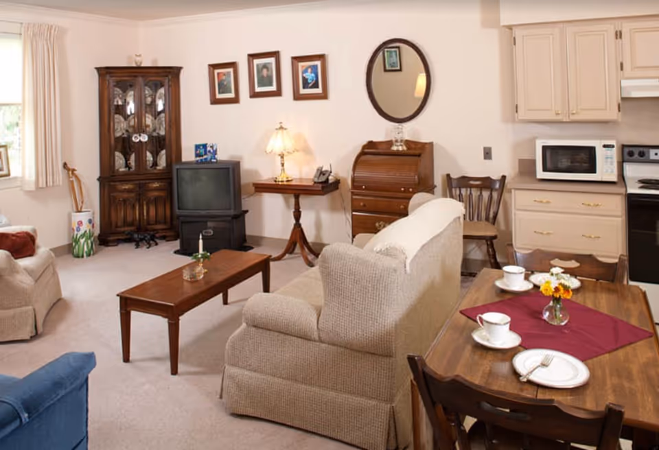 A cozy living area in a senior living facility featuring a beige sofa and armchair, a wooden coffee table with a candle, a wooden dining table set with cups, plates, and a small flower vase on a burgundy placemat. The room includes a wooden cabinet, an old-style TV, a small side table with a lamp, a wooden roll-top desk, and a kitchen area with a microwave and stove. The walls are decorated with framed pictures and an oval mirror.