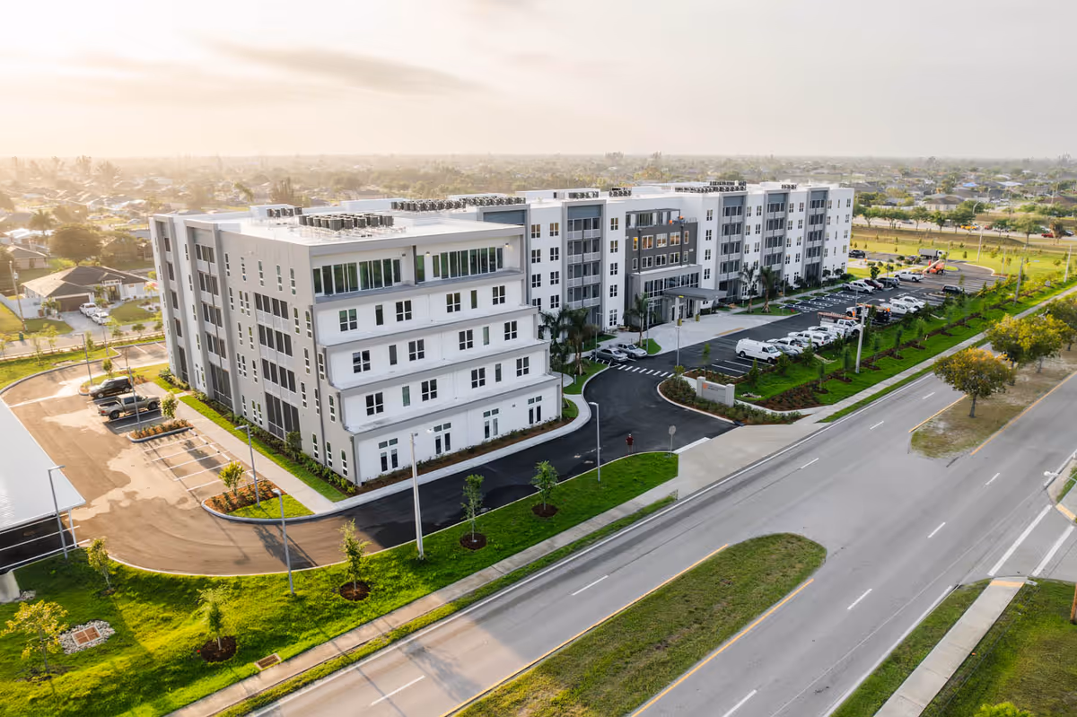 Aerial view of a modern multi-story senior living facility named The Gallery at Cape Coral, surrounded by landscaped greenery, parking lots, and adjacent roads under a bright sky.