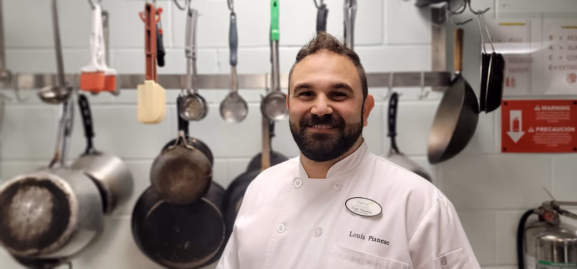 A smiling chef wearing a white uniform with the name tag 'Louis Pianese' stands in a kitchen with various pots, pans, and kitchen utensils hanging on the wall behind him.