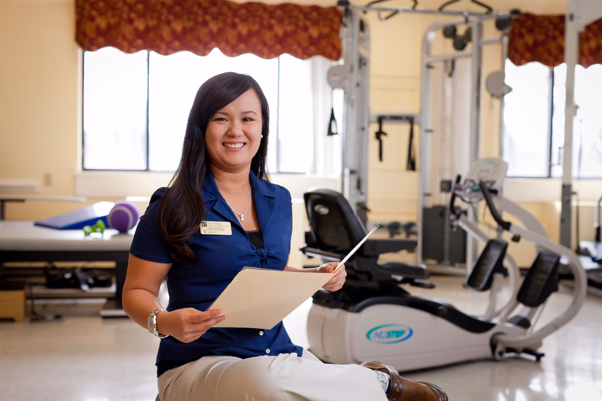 A smiling woman in a blue shirt holding a folder is seated in a rehabilitation or physical therapy room with exercise equipment and therapy tables in the background.