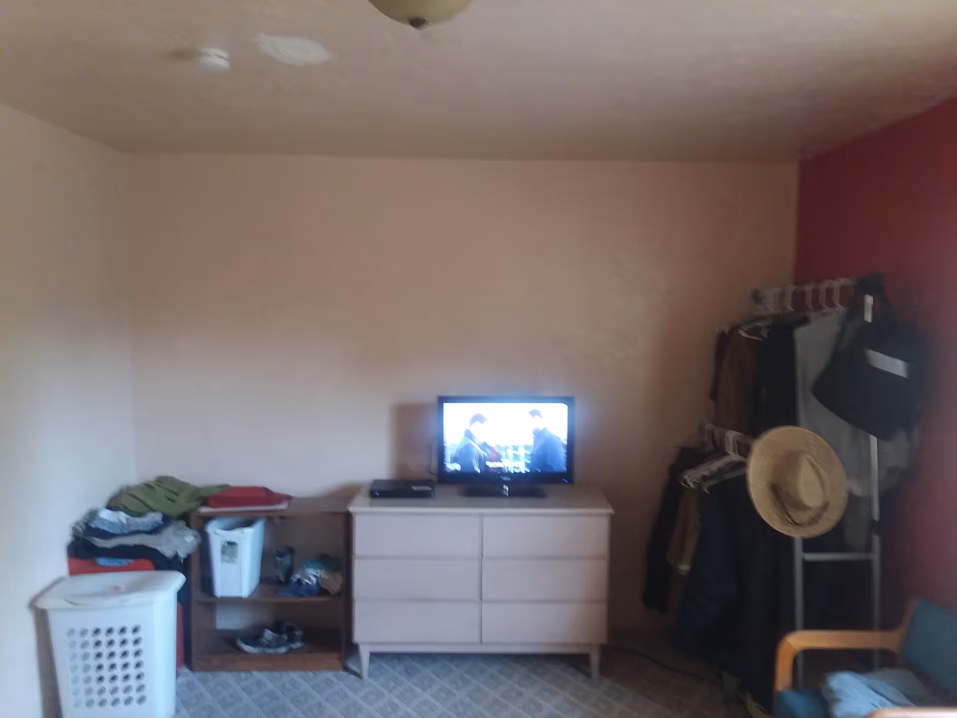 A small bedroom with a dresser holding a TV, a wooden shelf with folded clothes and shoes, a laundry basket, a clothing rack with hanging clothes and a straw hat, and a chair with clothes on it. The walls are painted light beige and red.