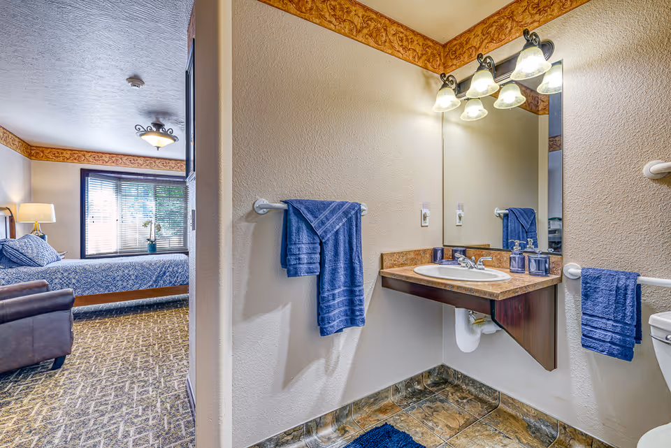 View of a senior living facility room showing a bedroom area with a bed, armchair, and window with blinds, adjacent to a bathroom area with a sink, mirror, toilet, and blue towels hanging on towel racks.