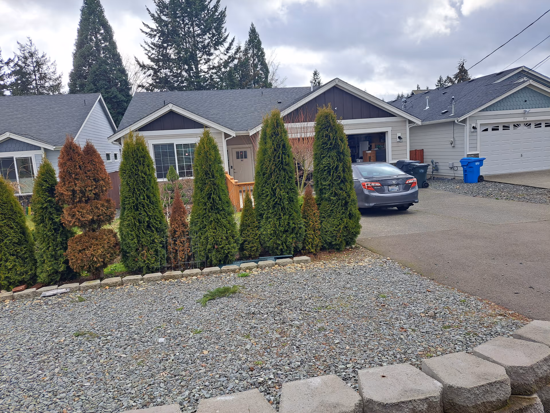 Front exterior view of a single-story house with a gray roof and beige siding, partially obscured by a row of tall evergreen shrubs. A gray car is parked in the driveway next to the house. The sky is cloudy and there are tall trees in the background.