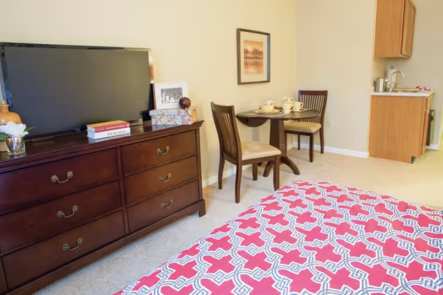 A cozy senior living bedroom featuring a bed with a red and white patterned bedspread, a wooden dresser with a flat-screen TV on top, and a small dining table with two chairs set against a beige wall. A framed picture hangs above the table, and a kitchenette area is visible in the background.