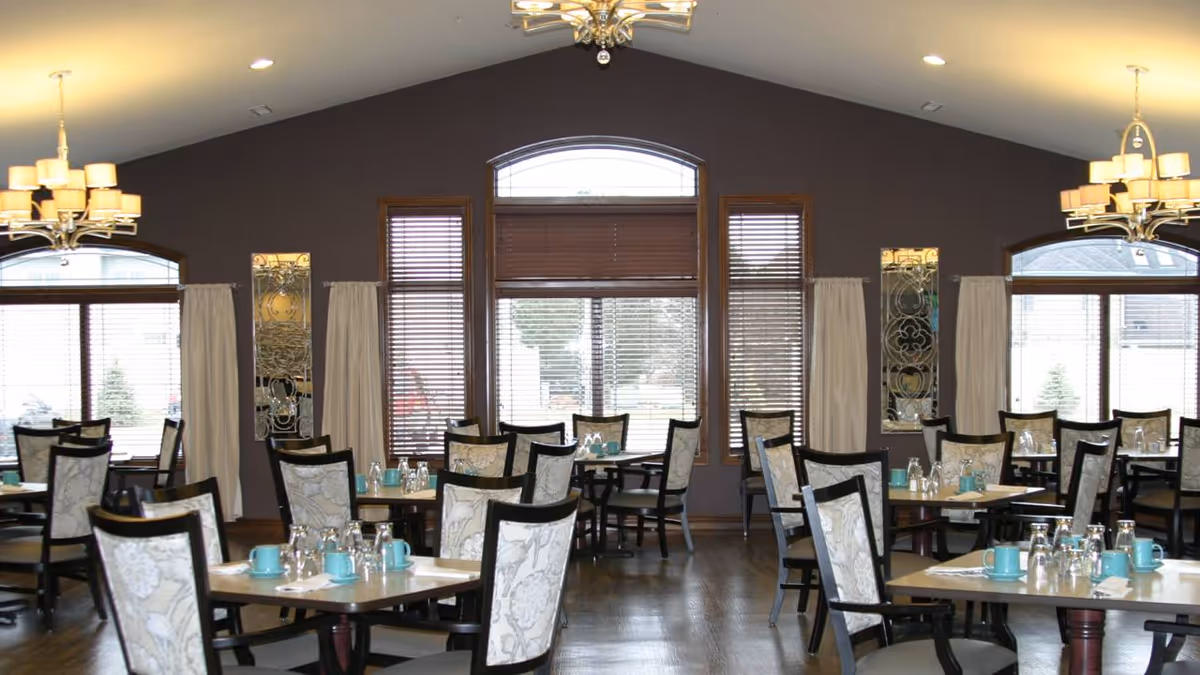 Spacious dining room with several set tables and cushioned chairs, large arched windows, and ceiling chandeliers.