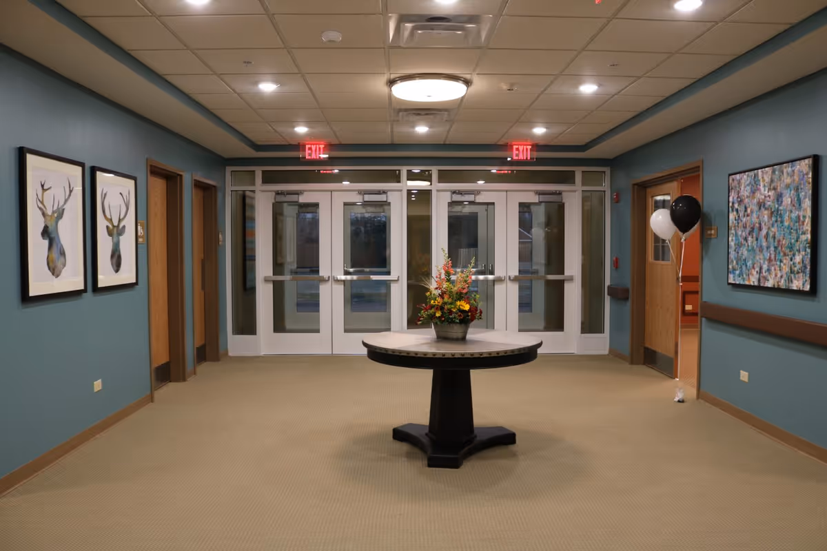 Lobby hallway with double glass entrance doors, a central round table topped with a flower arrangement, teal walls with framed art, and exit signs overhead.