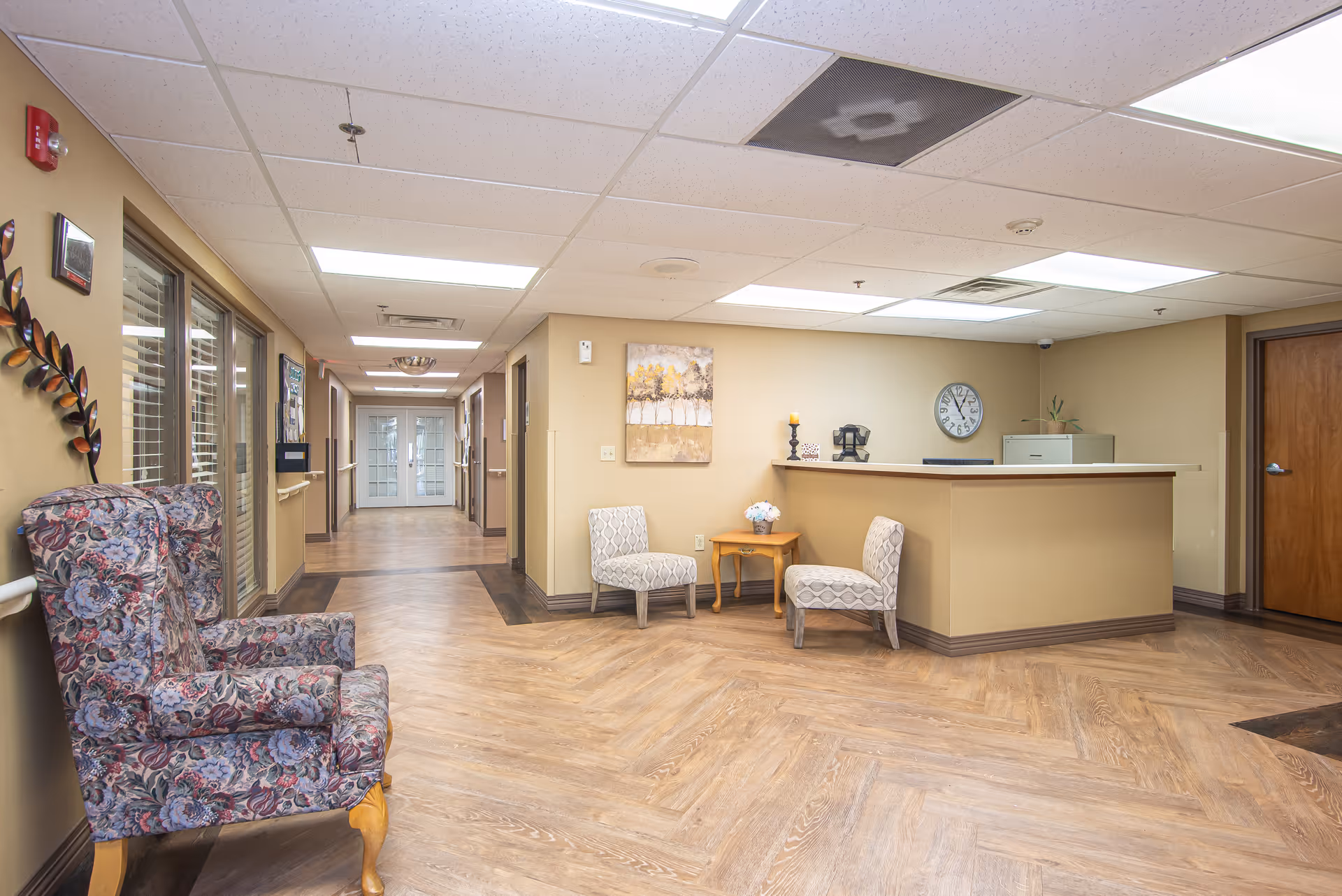 Interior view of a senior living facility hallway with a reception desk on the right side, two patterned chairs and a small wooden table with a flower vase. There is a floral armchair on the left side near windows with blinds. The hallway extends to double glass doors at the far end, with beige walls and wood-patterned flooring.