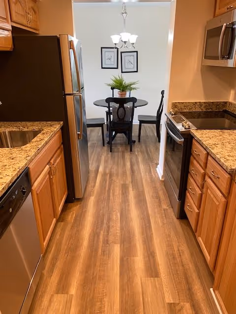 View down a kitchen with wooden cabinets, granite countertops, stainless steel appliances including a refrigerator, dishwasher, stove, and microwave. At the end of the kitchen is a small dining area with a round black table, four black chairs, a potted plant centerpiece, two framed pictures on the wall, and a chandelier overhead.