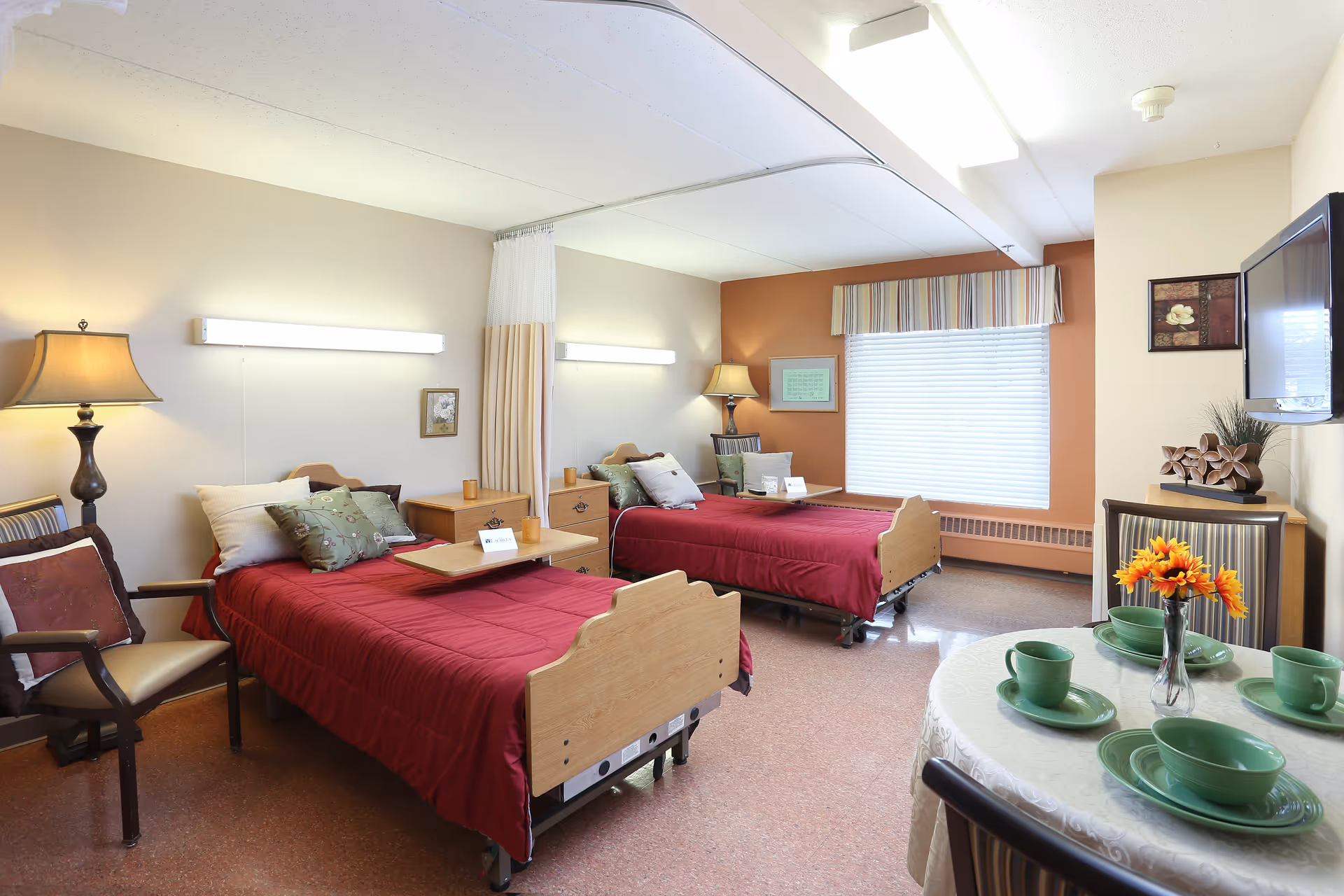 A bright and clean senior living bedroom with two hospital-style beds covered in red bedspreads, each with pillows and a bedside table. The room has a window with blinds and a striped valance, a round table set with green dishes and a vase of orange flowers, a wall-mounted TV, and comfortable chairs. The walls are painted in neutral tones with one accent wall in orange.