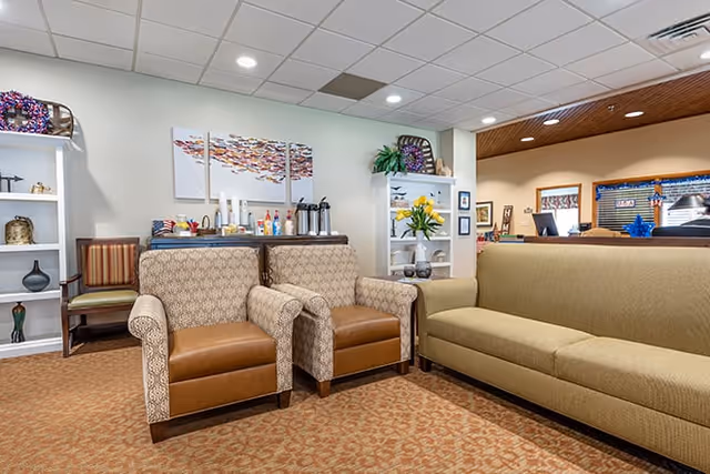 A comfortable senior living lounge with two armchairs, a sofa, decorative shelving and a refreshment station.