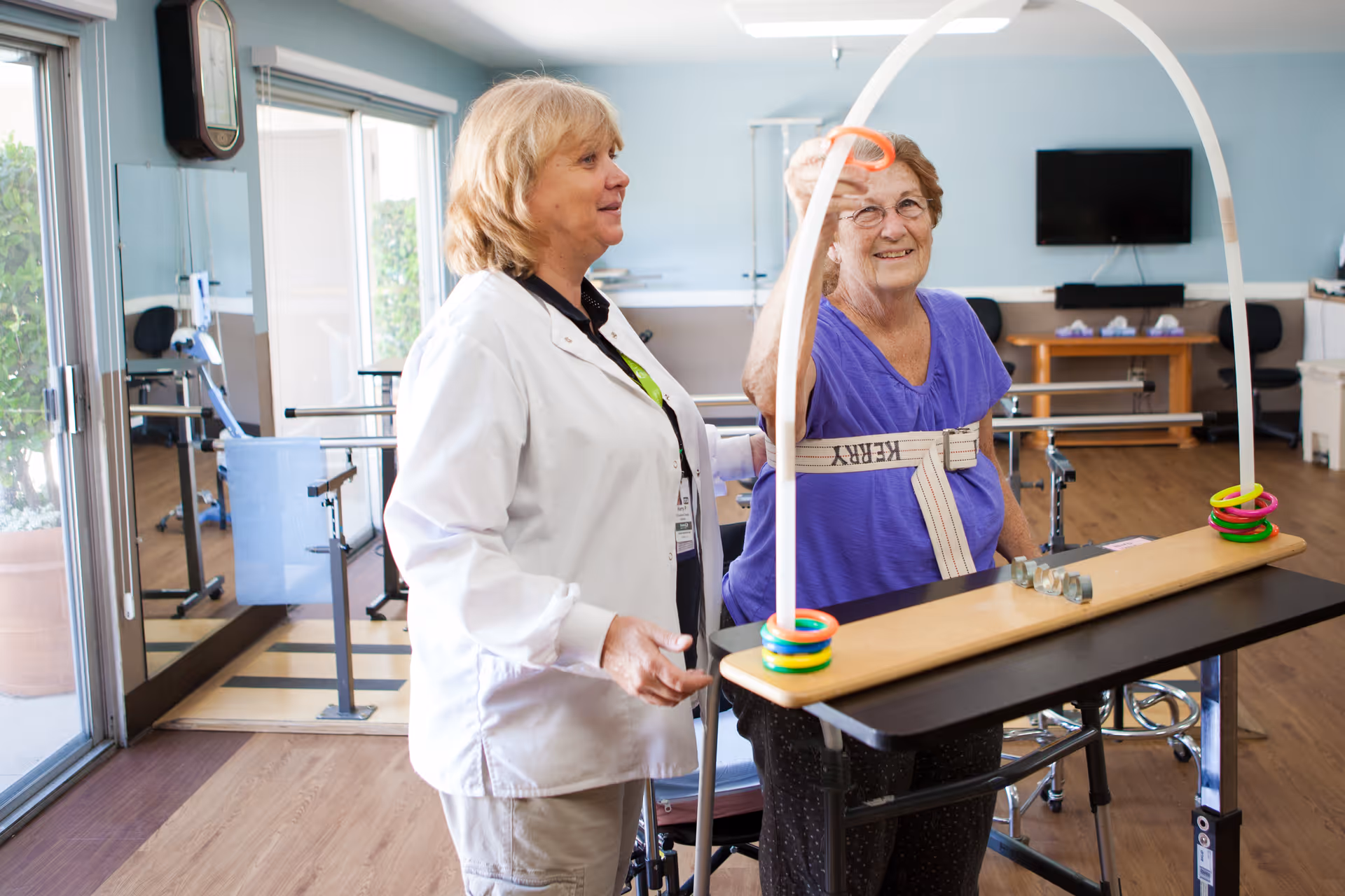 An elderly woman wearing a purple shirt is participating in a physical therapy exercise involving tossing rings onto a stand. A healthcare professional in a white coat is standing beside her, providing support and encouragement. The room has wooden floors, large windows, and exercise equipment in the background.