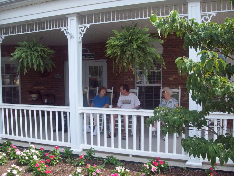 Three elderly people sitting and talking on rocking chairs on a covered porch with white railings and hanging ferns. There are flowers and greenery in front of the porch and a brick wall with windows and a door behind them.