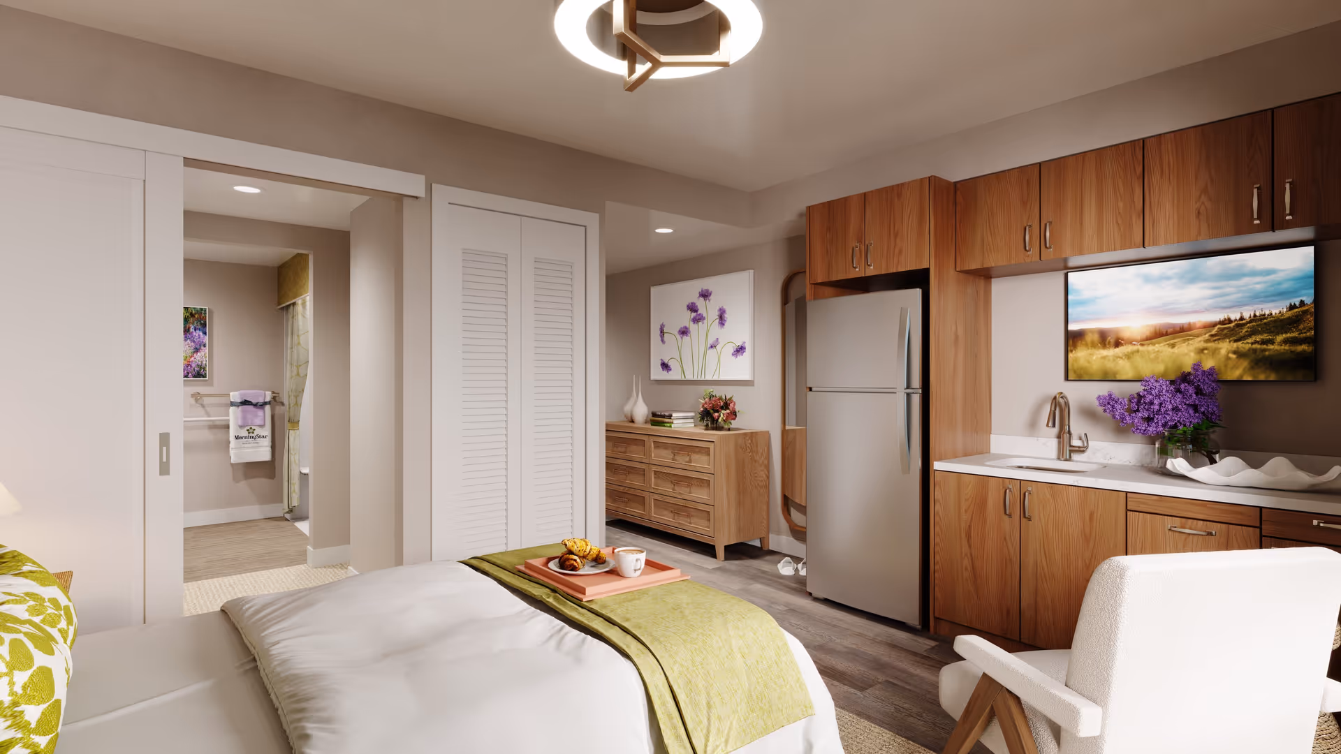 A cozy senior living studio apartment featuring a bed with green and white bedding, a tray with croissants and a cup of coffee, a wooden dresser with decorative items and flowers, a kitchenette with wooden cabinets, a refrigerator, a sink, and a wall-mounted TV displaying a scenic landscape. A white armchair is positioned near the kitchenette, and a bathroom is visible through an open sliding door.