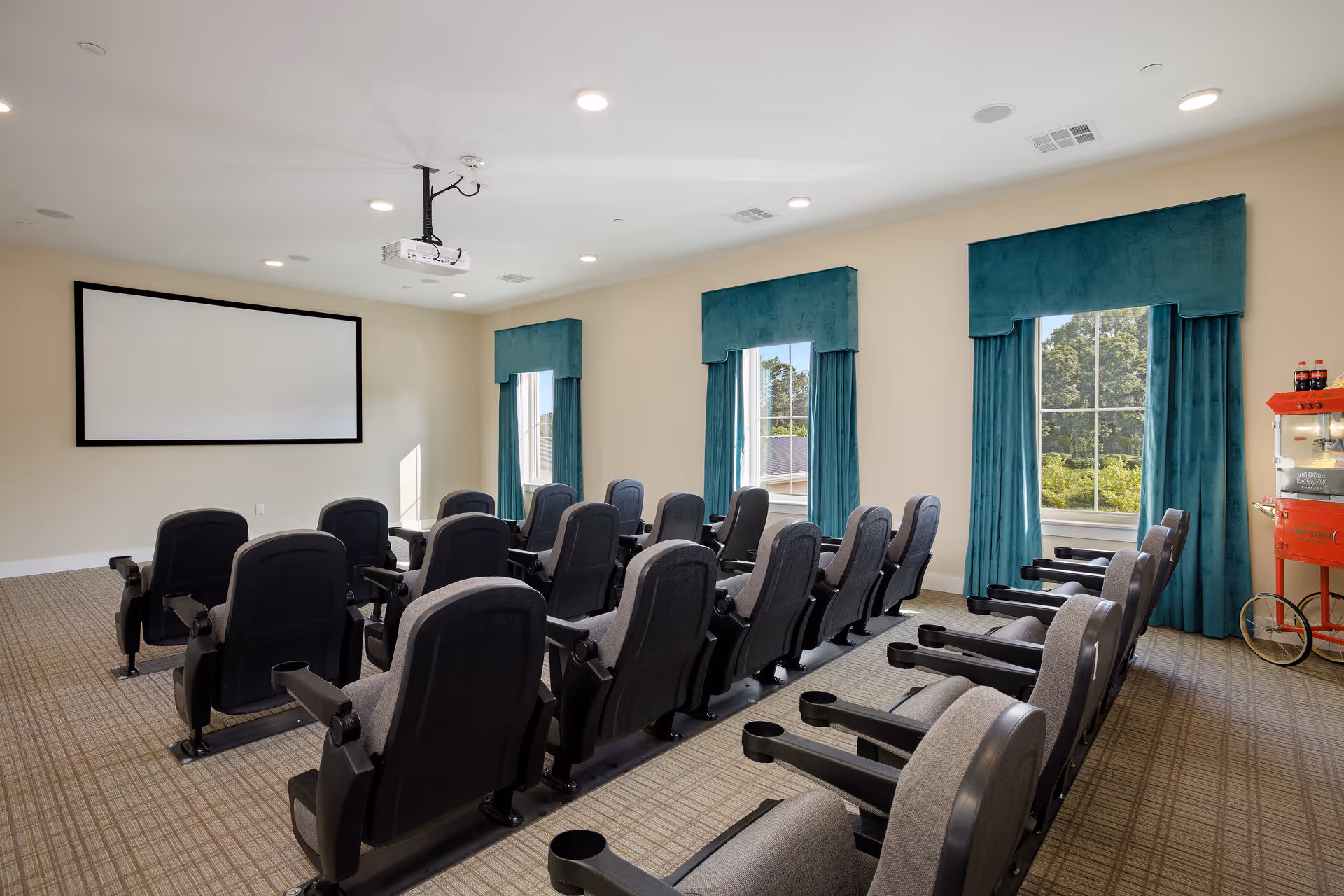 A small theater room with two rows of black and gray cushioned seats facing a large blank projection screen on the wall. The room has beige walls, three windows with teal curtains, a ceiling-mounted projector, and a red popcorn machine in the corner.