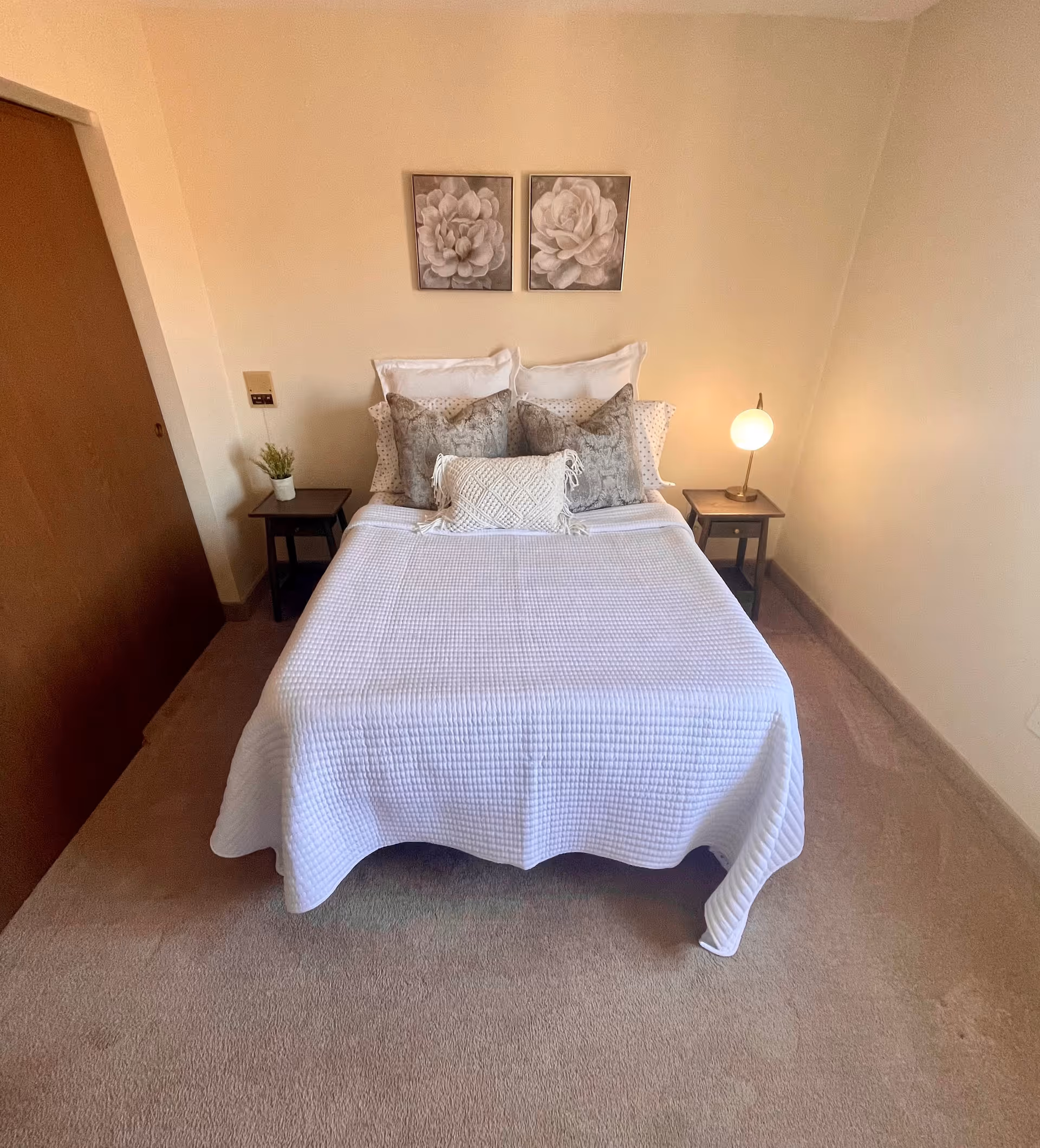 A neatly made bed with a white textured bedspread and multiple pillows in a small bedroom. There are two framed floral artworks hanging above the bed. On each side of the bed, there are small dark wooden nightstands; the left one has a small potted plant and the right one has a round lamp emitting warm light. The room has beige walls and carpeted floor.