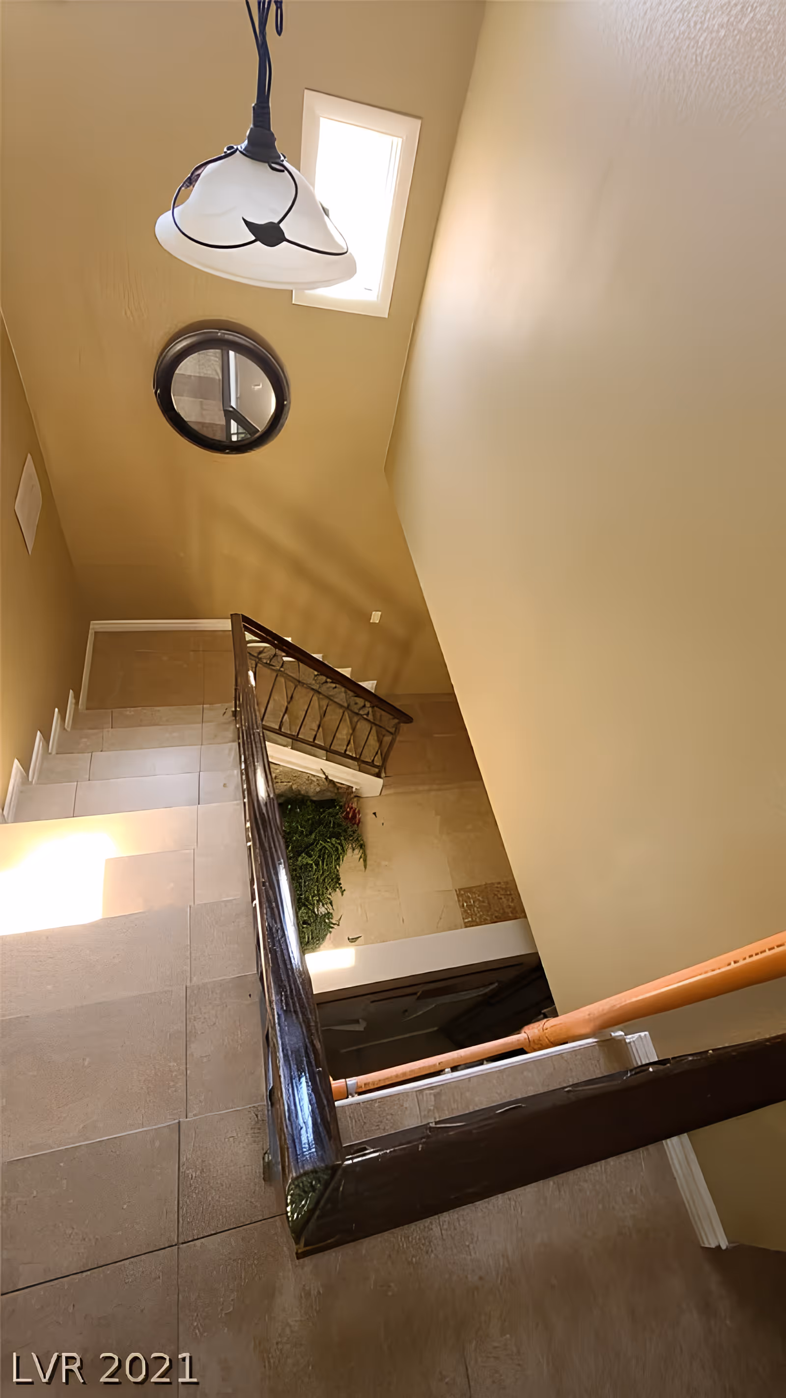 View down an interior stairwell with tiled steps, a wooden railing, hanging light fixture and round mirror on beige walls.