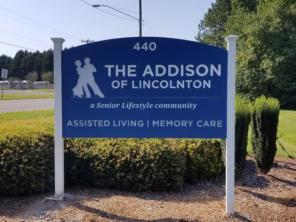 A blue and white outdoor sign for The Addison of Lincolnton, a senior lifestyle community offering assisted living and memory care, located at 440. The sign features a silhouette of two people dancing and is surrounded by greenery and trees.