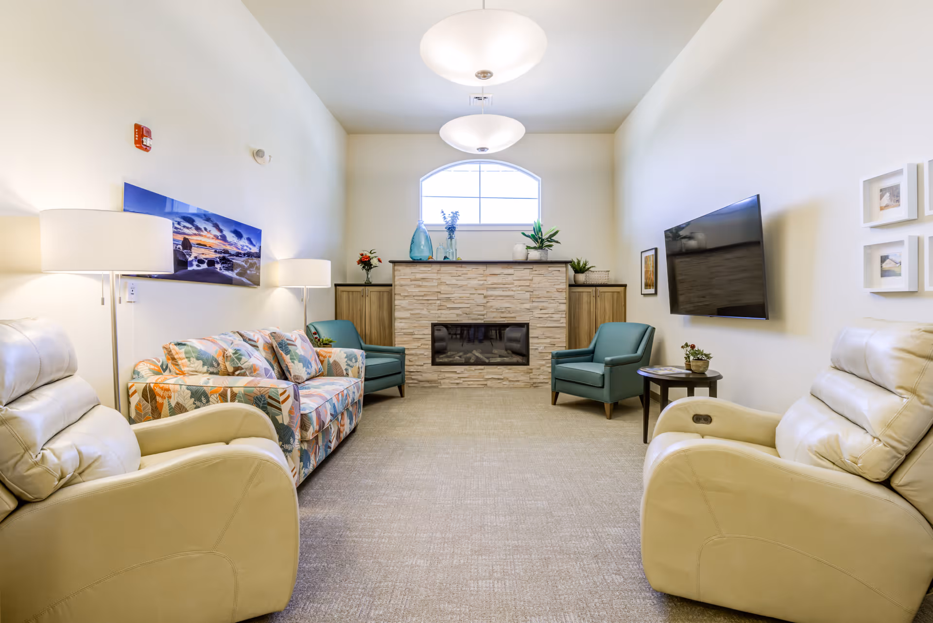 A bright and cozy living room with two beige recliners, a floral patterned sofa, two teal armchairs, a stone fireplace with decorative vases and plants on the mantel, a wall-mounted flat-screen TV, and soft lighting from ceiling fixtures and floor lamps.