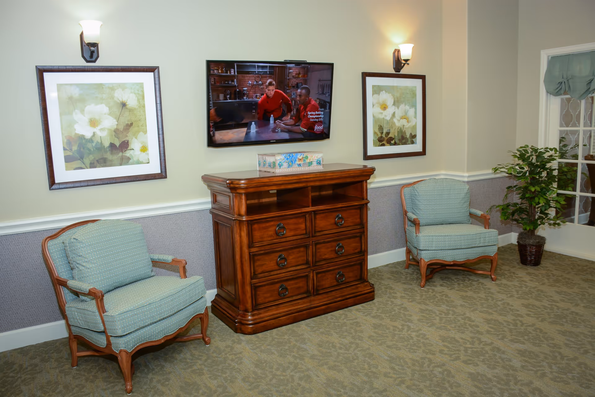 A cozy sitting area with two light blue cushioned armchairs with wooden frames placed on either side of a wooden cabinet with drawers. Above the cabinet is a wall-mounted flat-screen TV showing a cooking show. Two framed floral paintings hang on the wall, each above one armchair. A potted plant is placed near a glass door with a green curtain. The room has soft lighting from two wall sconces.