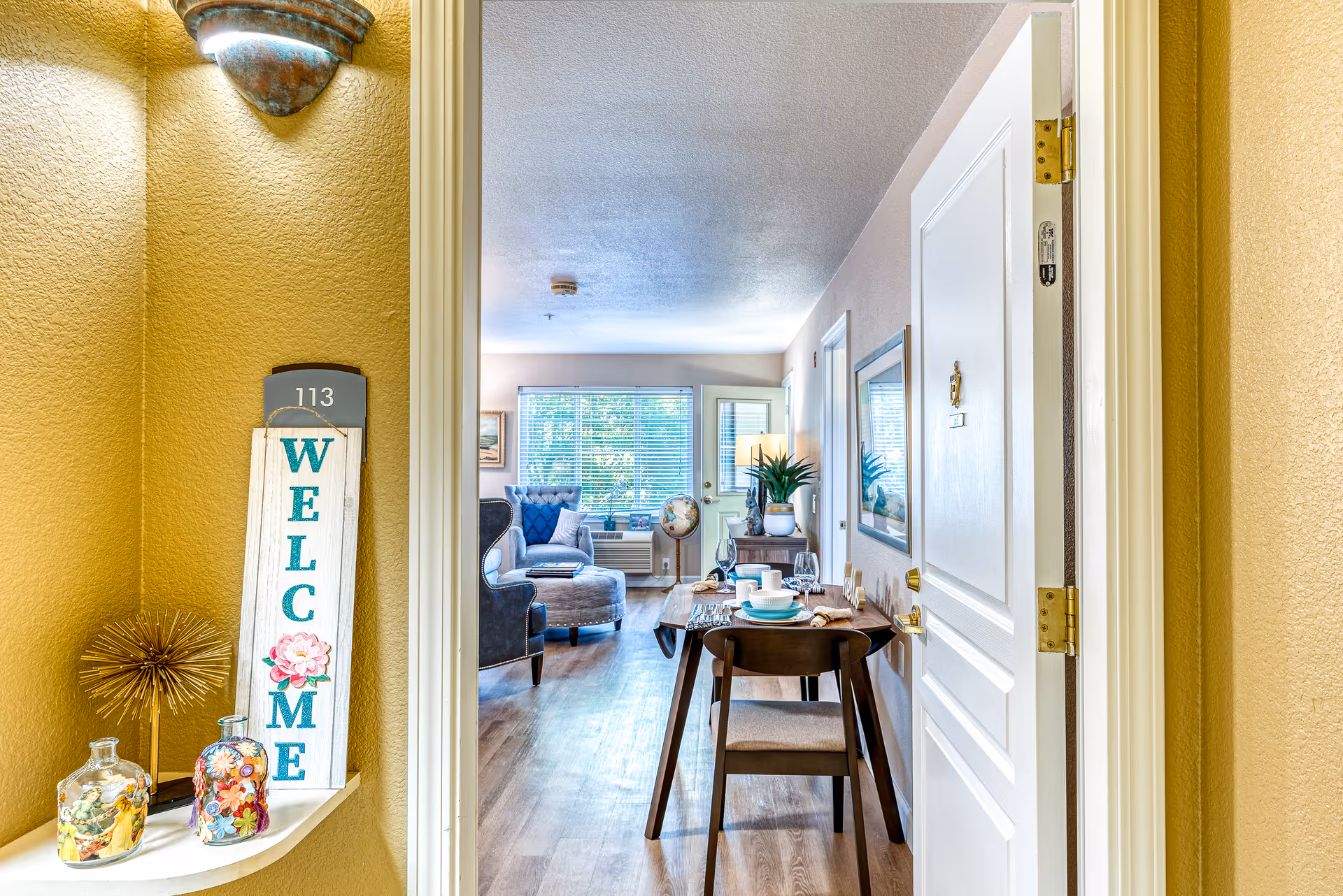 View through an open door into a bright and cozy senior living apartment featuring a small dining table set for two, a comfortable armchair, a window with blinds, and decorative items including a globe and plants. A welcome sign and decorative jars are placed on a small ledge near the door marked 113.