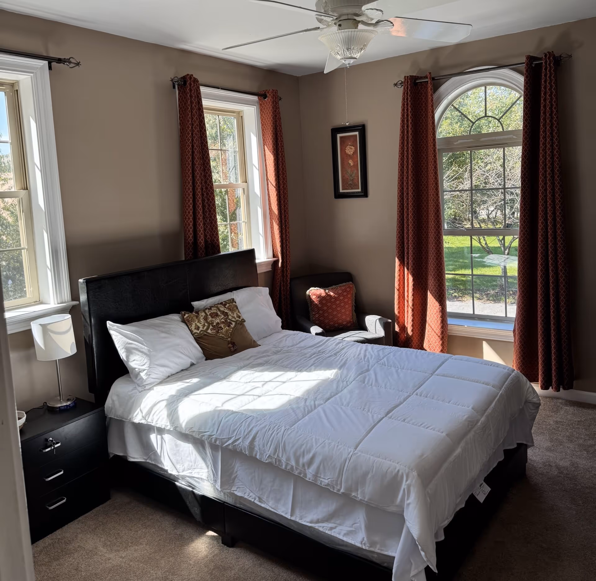 A bedroom with a large bed, nightstands, red curtains, and a ceiling fan.