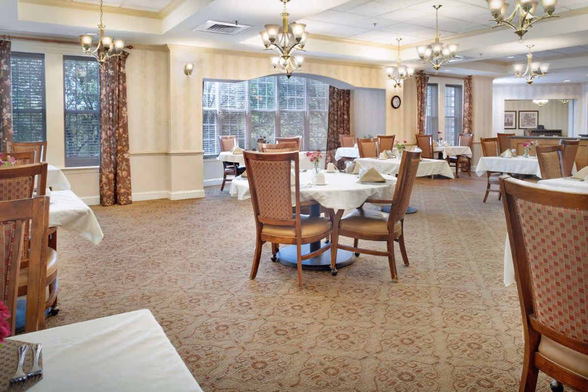 A spacious dining room with multiple round and rectangular tables covered with white tablecloths, each set with napkins and small flower vases. The room features patterned carpet, large windows with floral curtains, and elegant chandeliers hanging from the ceiling.
