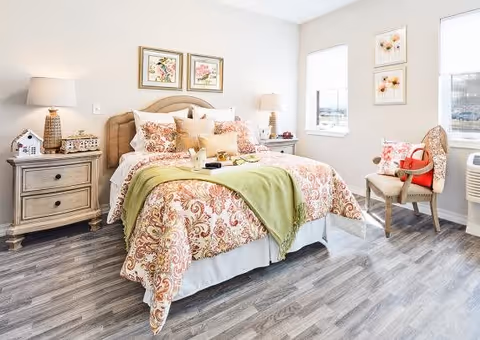 A bright and cozy bedroom with a large bed featuring a patterned bedspread and multiple pillows. There are two bedside tables with lamps, decorative framed pictures on the walls, a chair with a red handbag, and two windows letting in natural light. The floor has a wood-like finish.