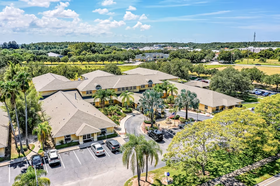 Aerial view of a senior living facility with multiple connected beige buildings surrounded by palm trees and greenery. There is a circular driveway with parked cars and a small landscaped area with an American flag in front of the main entrance. The background shows more trees, open fields, and a partly cloudy sky.