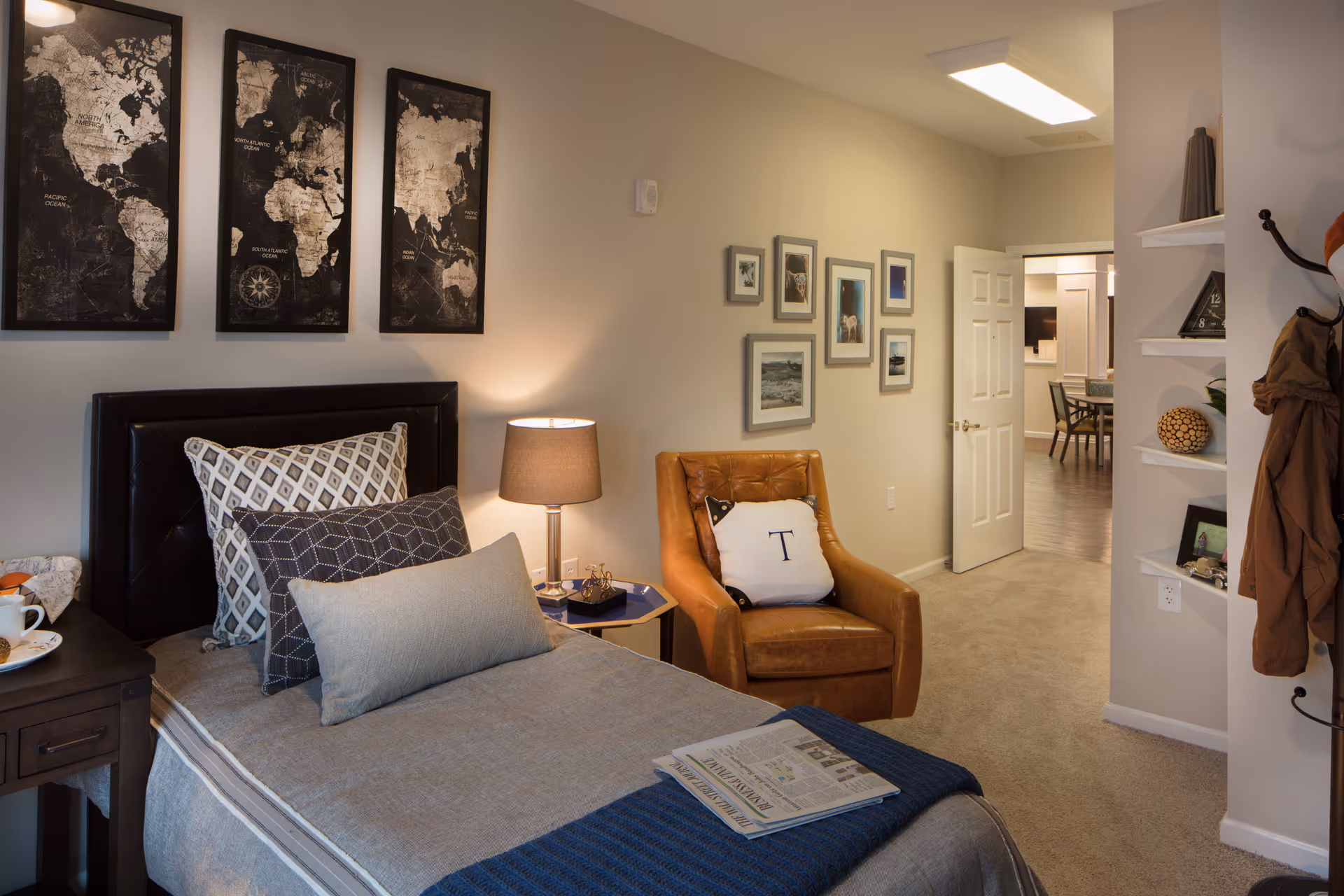 A cozy bedroom in an assisted living facility featuring a single bed with multiple pillows and a folded newspaper on a blue blanket. Next to the bed is a nightstand with a lamp and a cup. On the wall above the bed are three framed world map prints. A brown leather armchair with a white pillow marked with the letter 'T' is positioned near a wall adorned with several framed pictures. The room has beige walls and carpet, with an open door leading to another room with a dining table and chairs.