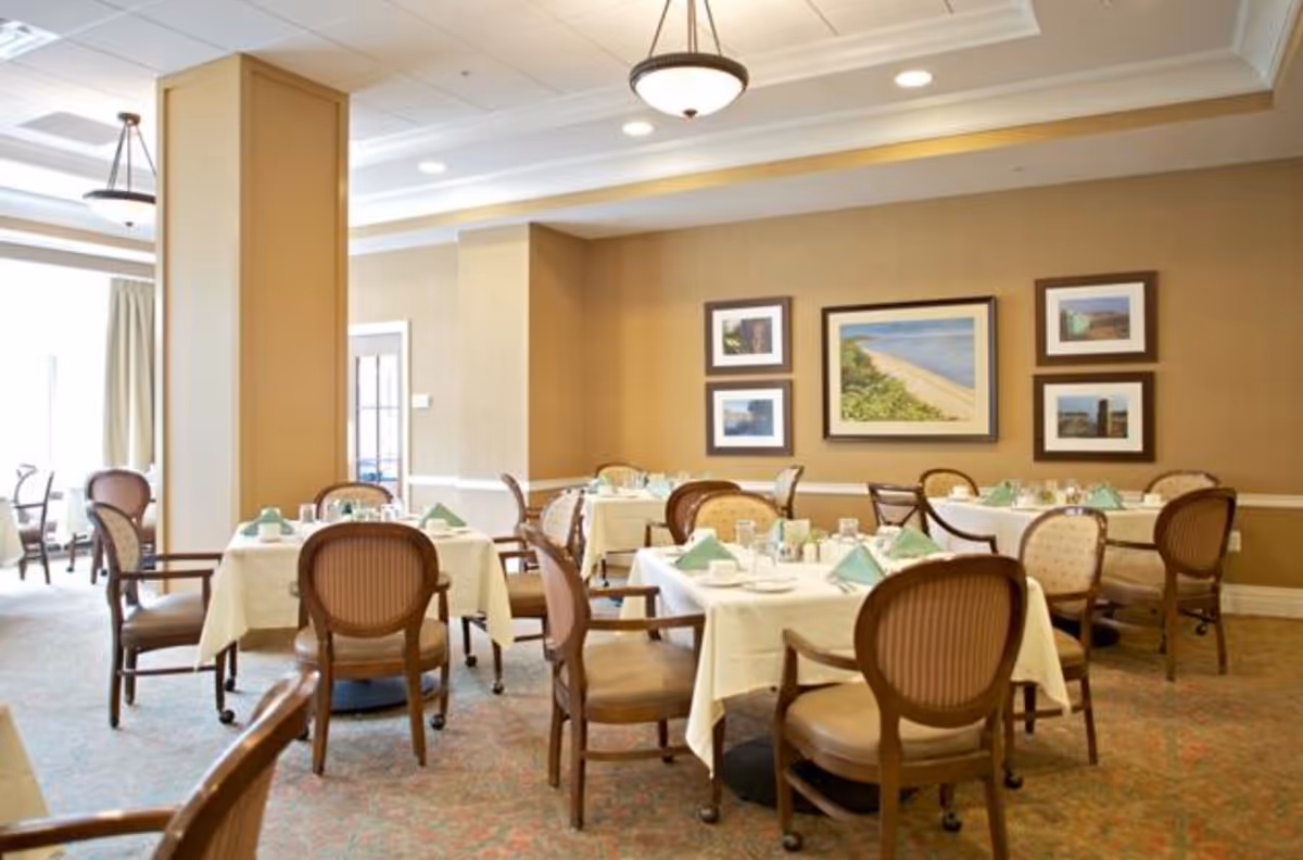 A dining room with several tables covered in white tablecloths, each set with plates, glasses, and green folded napkins. The room has beige walls adorned with framed pictures, and wooden chairs with cushioned seats surround the tables. The space is well-lit with ceiling lights and natural light from windows.