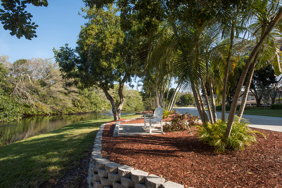 A peaceful outdoor area at Freedom Village of Bradenton featuring a small paved seating area with a white chair surrounded by palm trees and other greenery. A calm waterway runs alongside a grassy bank, and a paved road is visible in the background under a clear blue sky.
