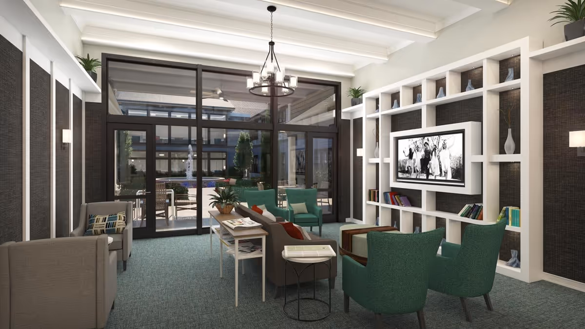 A modern living room with green armchairs, a gray sofa, and a white shelving unit with decorative items and books. A flat-screen TV is mounted on the shelving unit. Large glass doors lead to an outdoor courtyard with seating and greenery. The room has a chandelier and wall sconces providing lighting.