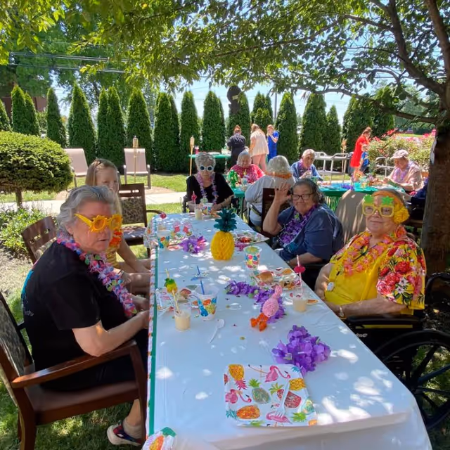 A group of elderly people sitting around a long outdoor table decorated with tropical-themed items such as a pineapple centerpiece, colorful leis, and festive cups. Some individuals are wearing bright, playful accessories like flower-shaped glasses and leis. The setting is a sunny garden area with green bushes and trees providing shade.
