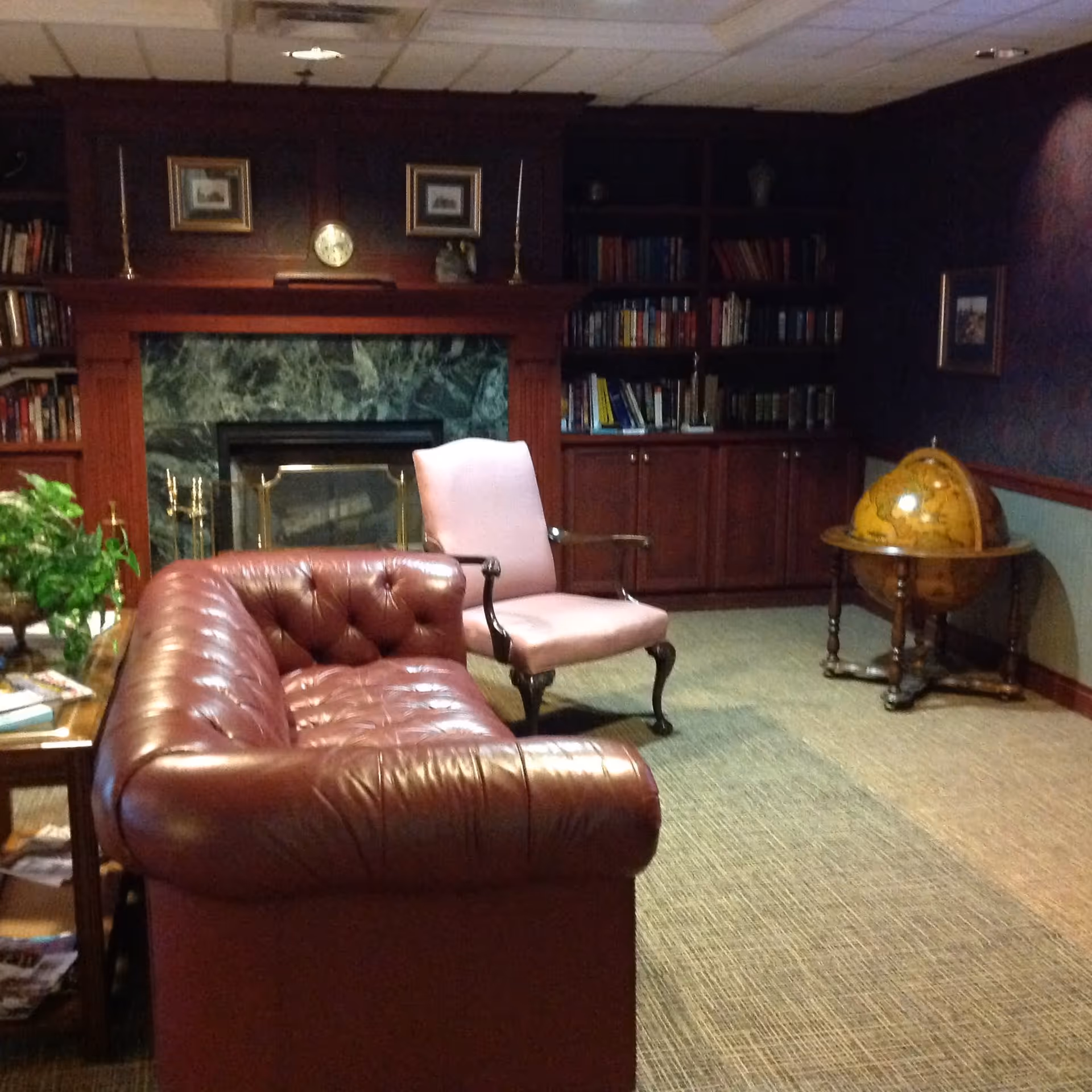 A cozy living room with a dark red leather tufted sofa, a light pink upholstered armchair, a wooden coffee table with plants and magazines, a fireplace with a green marble surround, built-in wooden bookshelves filled with books, and a vintage globe on a wooden stand.