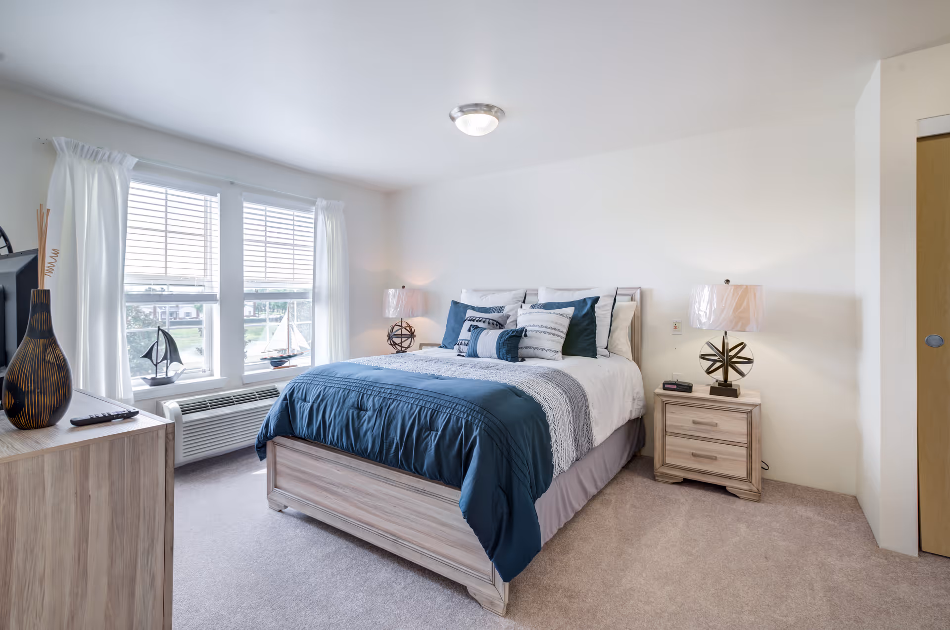 A bright and neatly arranged bedroom with a large bed featuring blue and white bedding and multiple pillows. There are two matching wooden nightstands on either side of the bed, each with a decorative lamp. A window with white curtains lets in natural light, and a wooden dresser with decorative items is visible on the left side of the room.