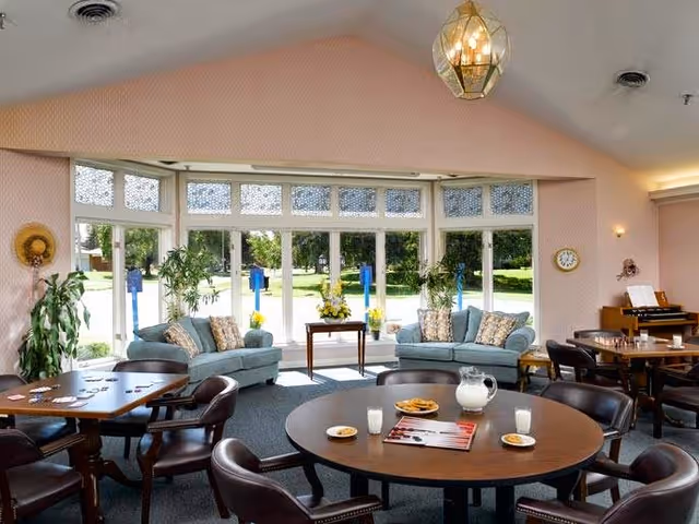A bright and spacious common area with large windows letting in natural light. The room features two light blue sofas with patterned cushions, several round tables with chairs around them, and a pitcher of milk with glasses and cookies on one table. There are plants near the windows and a piano in the corner, creating a welcoming and comfortable atmosphere.