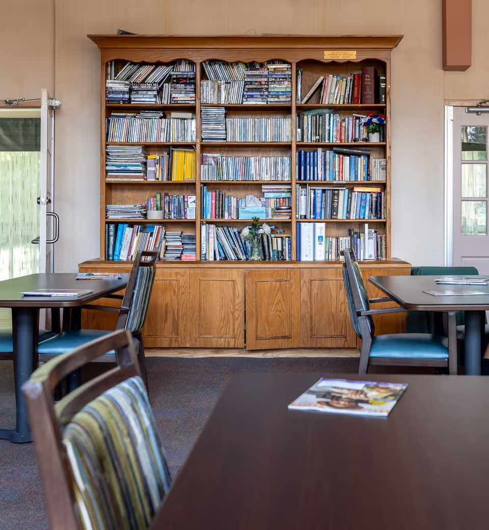 Interior view of a room with wooden bookshelves filled with books and DVDs. There are tables and chairs with striped cushions arranged in front of the shelves. The room has a carpeted floor and doors with glass panels on either side of the bookshelf.