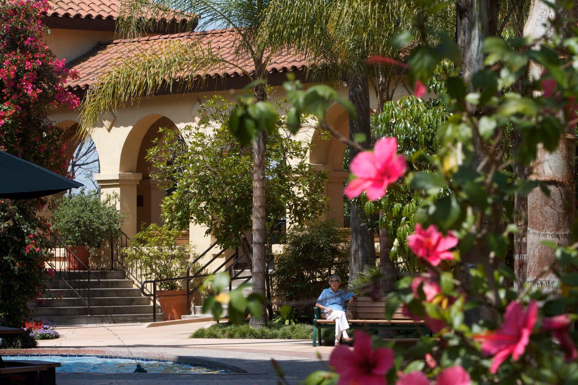 An elderly person sitting on a bench in a garden area with vibrant pink flowers in the foreground. The background shows a building with arched doorways and a tiled roof, surrounded by lush greenery and palm trees.