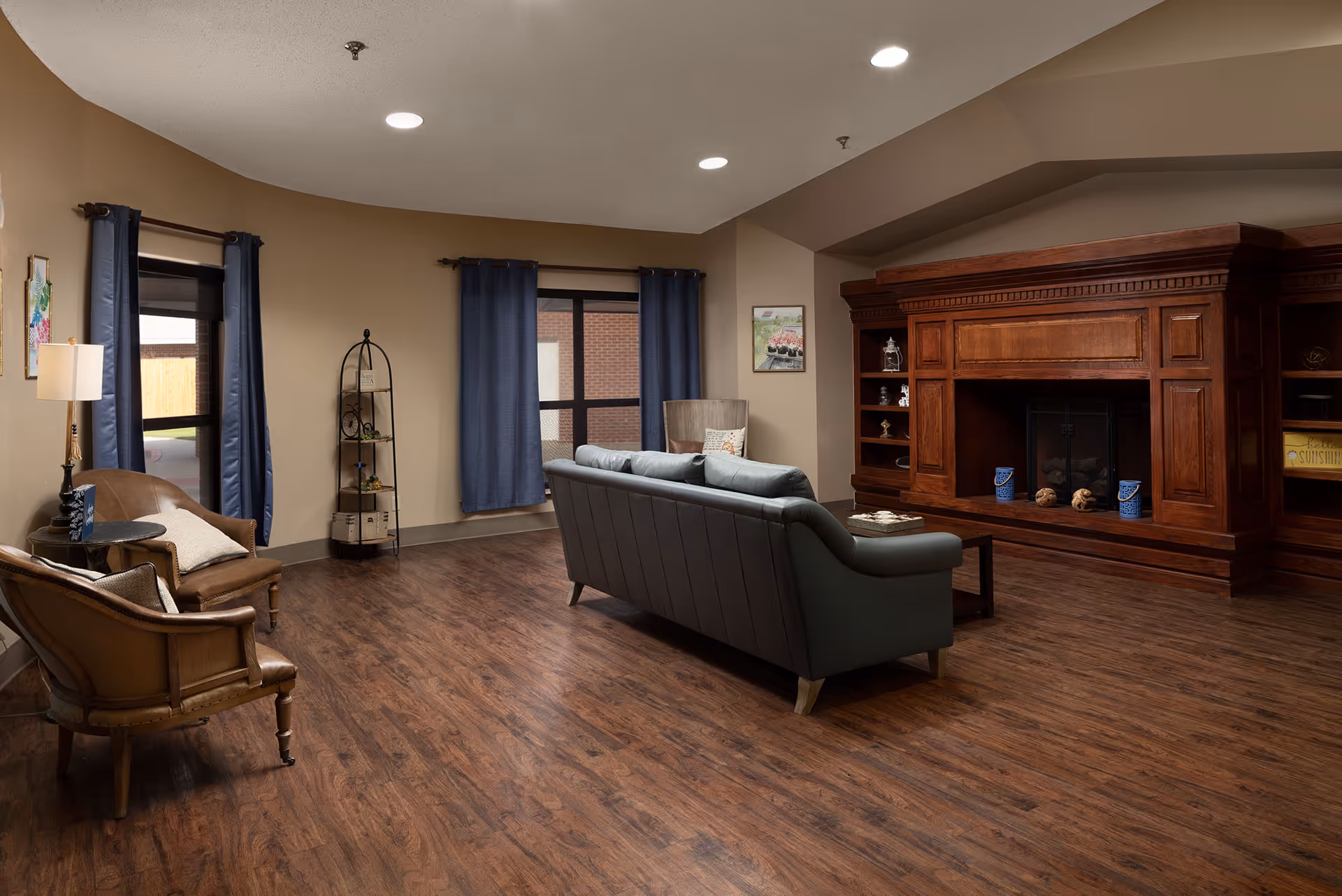 A cozy living room area with wooden flooring, a large wooden fireplace, a gray sofa facing the fireplace, two brown armchairs with cushions, a small round side table with a lamp, blue curtains on windows, and a decorative shelving unit in the corner.