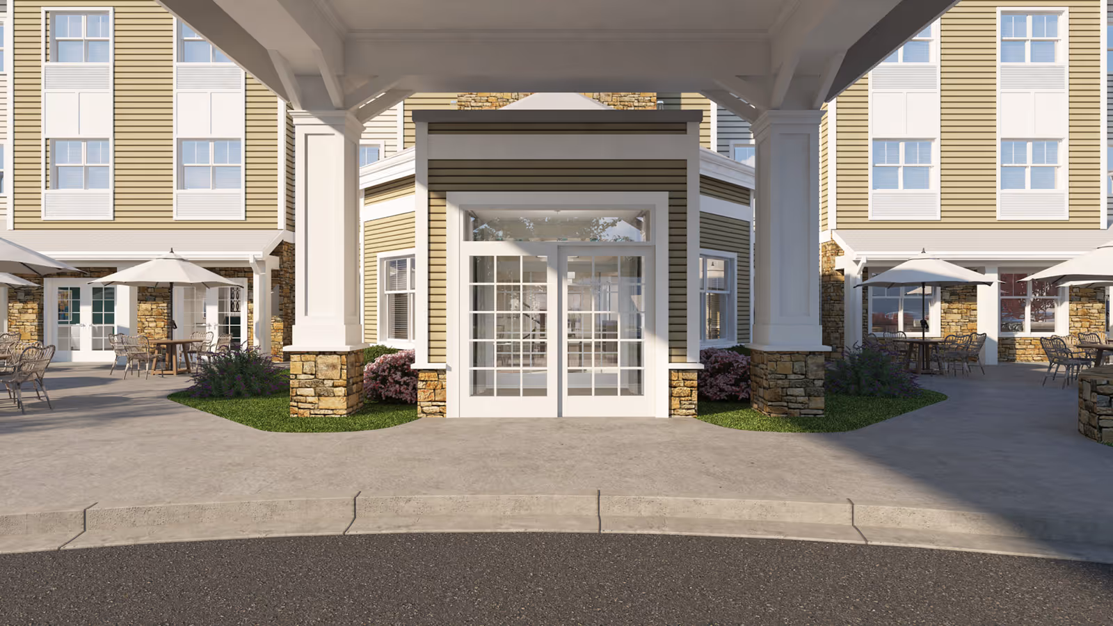 Entrance area of a senior living facility with a covered drop-off zone supported by white columns with stone bases. The building exterior features yellow siding with white trim and multiple windows. There are outdoor seating areas with tables, chairs, and umbrellas on either side of the entrance, along with landscaped greenery and flowers.