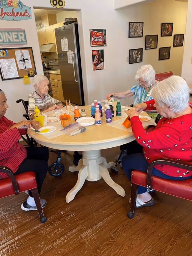 Four elderly women seated around a round table engaged in a painting activity with various bottles of paint and brushes on the table. The room has a kitchen area in the background and walls decorated with vintage-style signs and framed pictures.