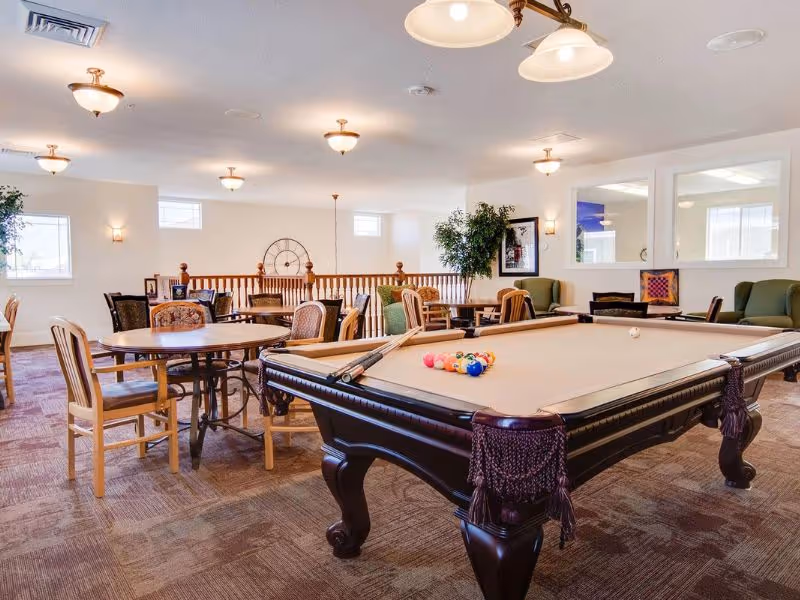 A spacious senior living common area featuring a pool table with billiard balls and cues, several round tables with chairs, comfortable armchairs, and decorative plants. The room is well-lit with ceiling lights and has windows allowing natural light.