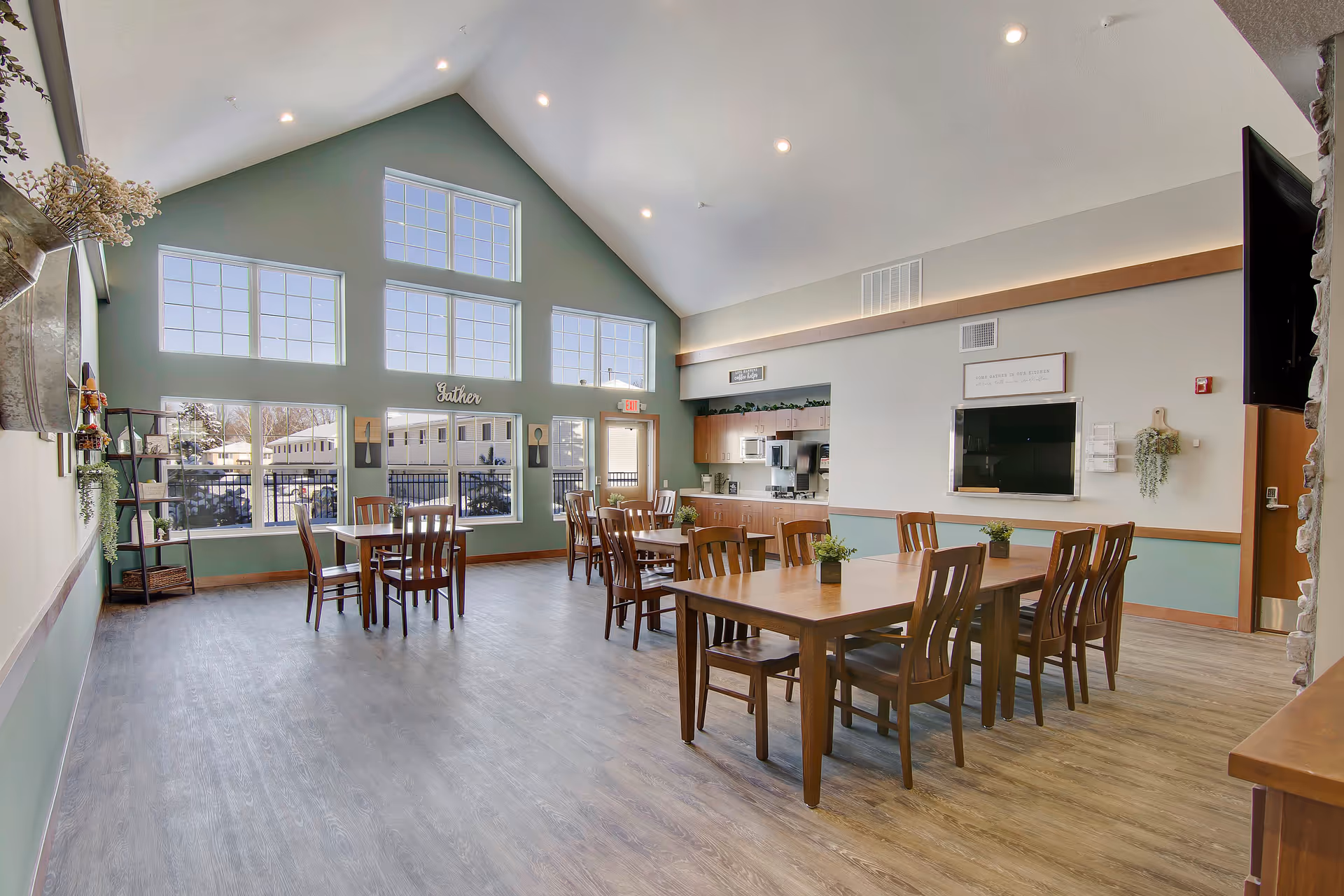 A spacious, well-lit dining room with large windows allowing natural light to fill the space. The room features multiple wooden tables and chairs arranged neatly, small green plants as centerpieces, and a kitchen area with cabinets and appliances in the background. The walls are painted in soft green and white tones, and there is a sign that says 'Gather' above the windows.