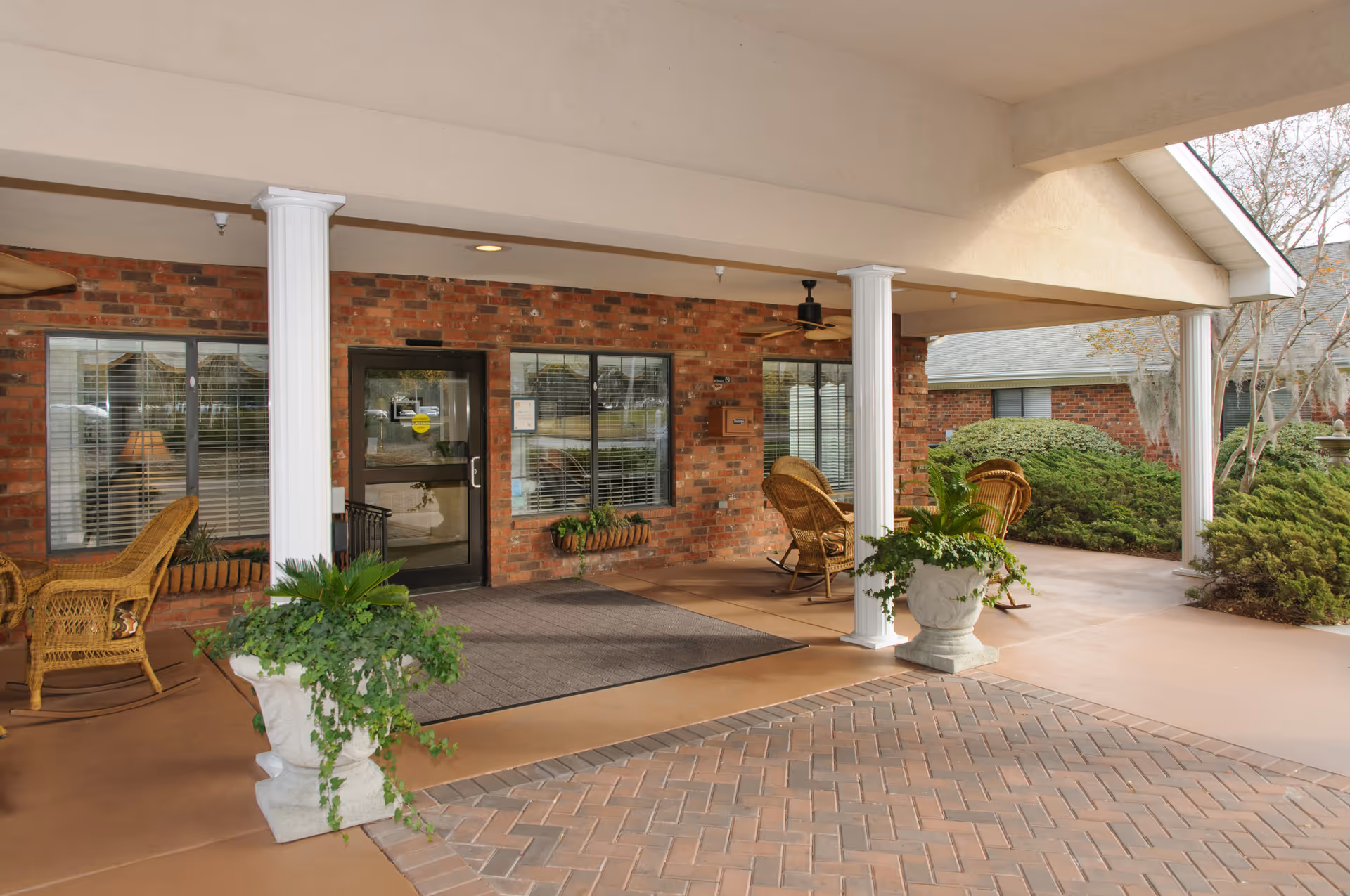 Covered entrance area of a brick building with white columns, wicker chairs, potted plants, and a glass door leading inside. There are windows with blinds on either side of the door and some greenery around the building.