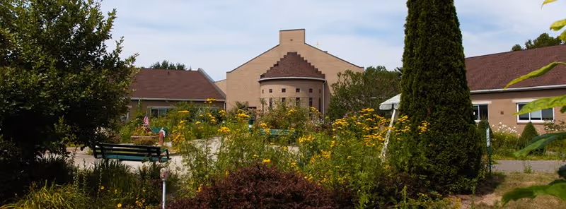 Front exterior of a brick senior living building with a landscaped courtyard, benches, and flowering plants.